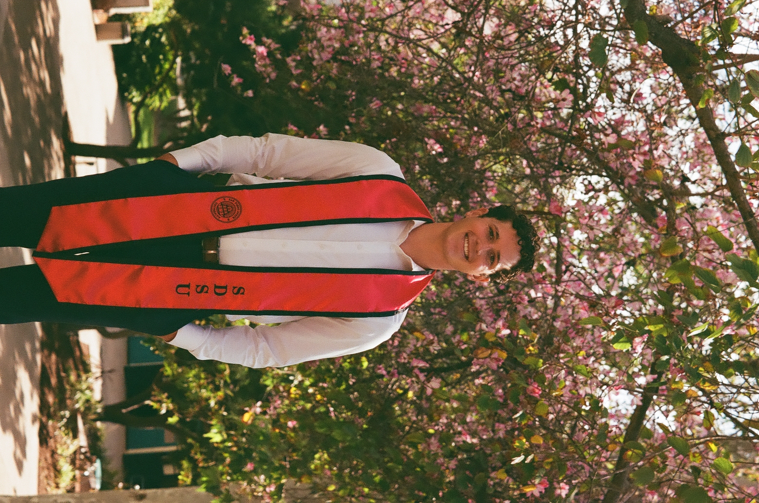 Owen smiling in SDSU graduation sash under a pink flowering tree