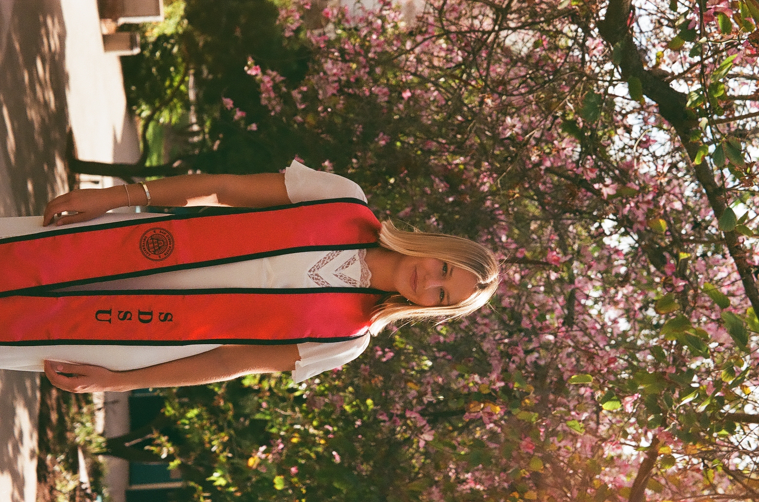 Anabelle smiling in SDSU graduation sash under a pink flowering tree