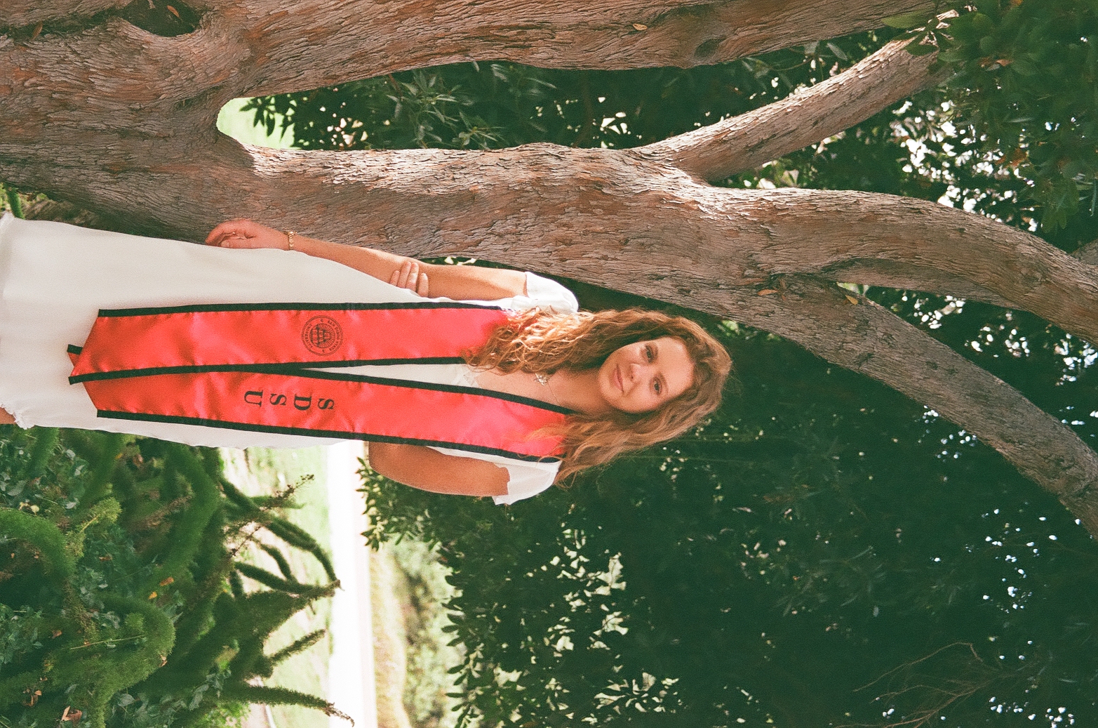 Kiera smiling in SDSU graduation sash leaning against a tree