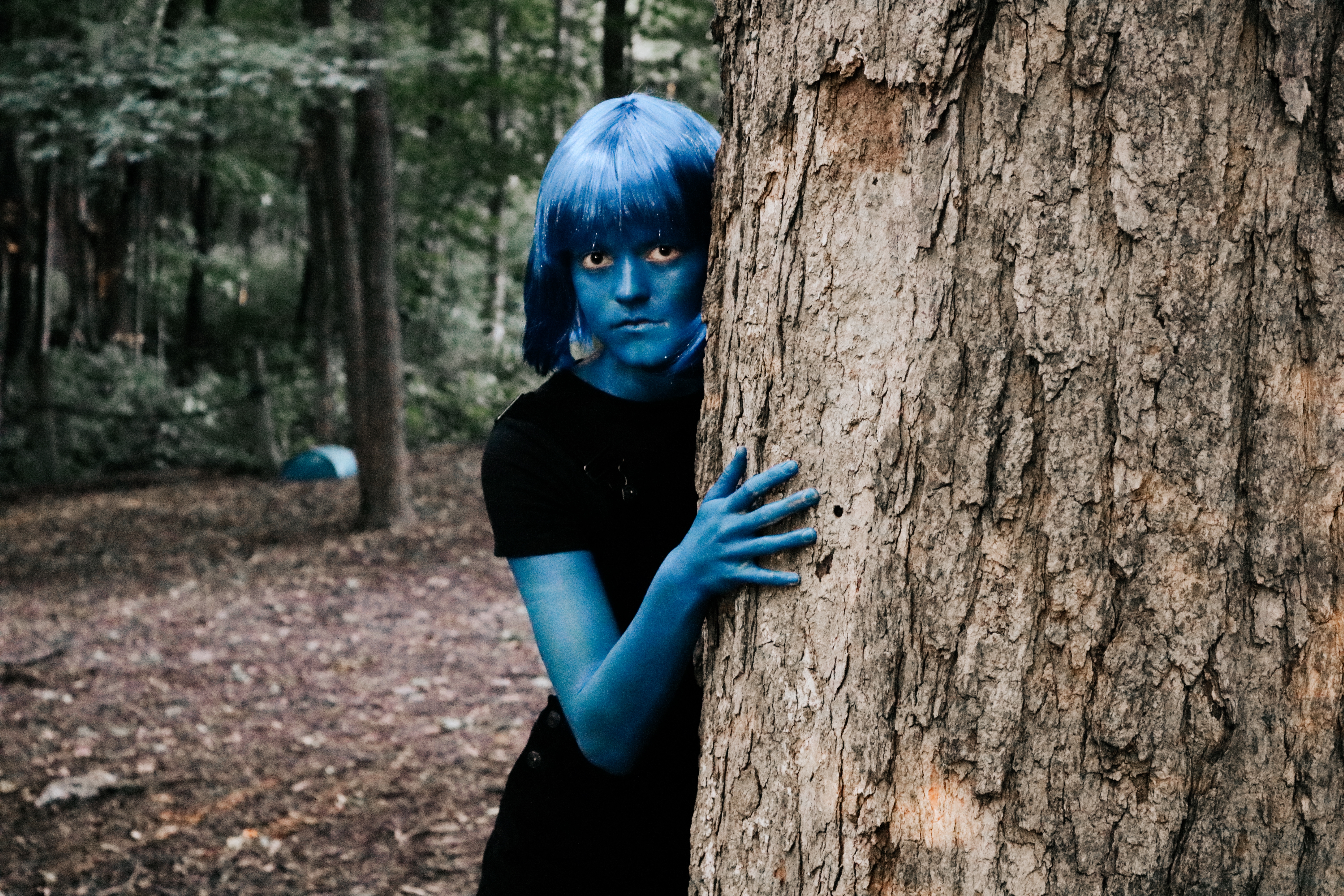 Figure with blue body paint and wig peering around a tree in the forest