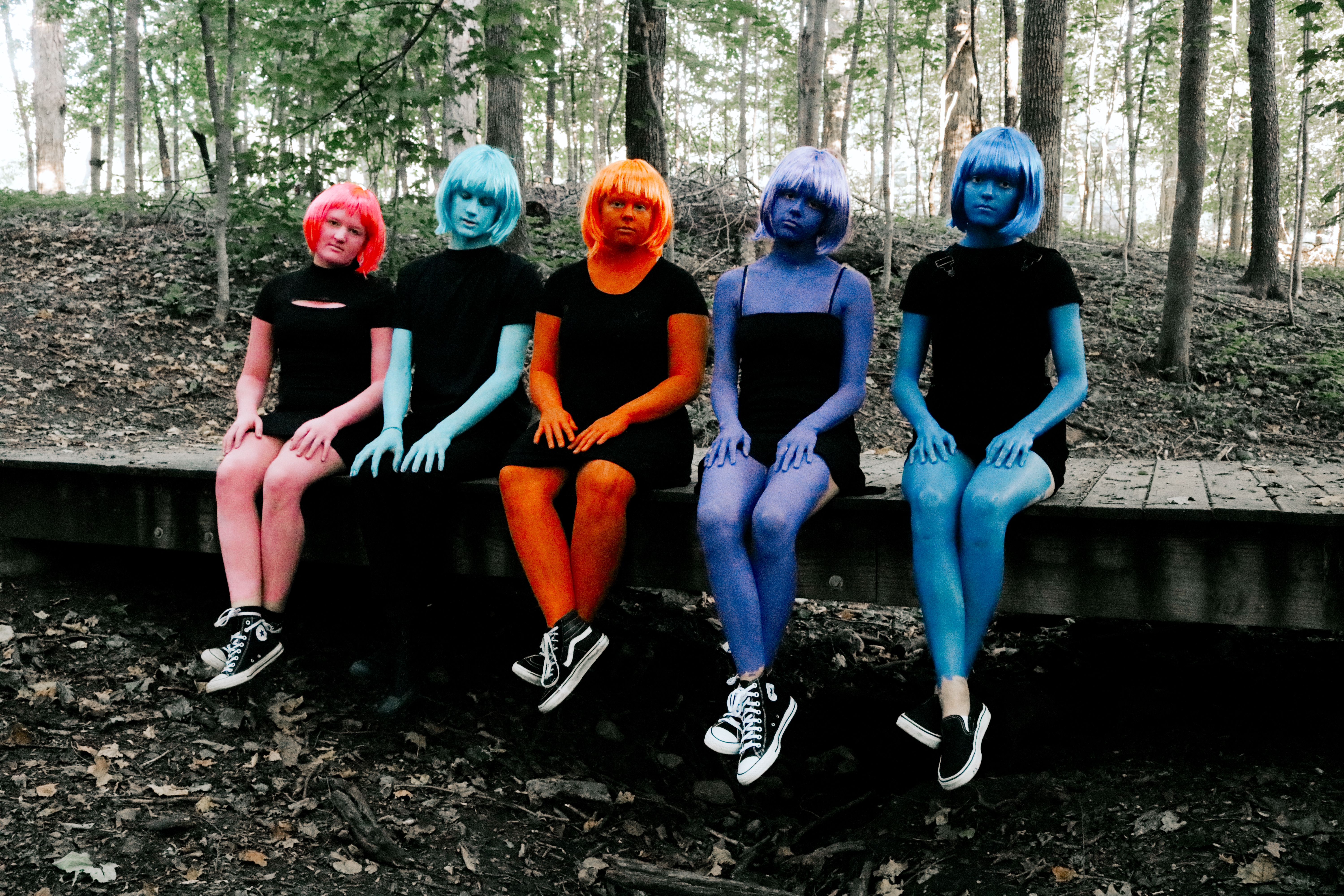 Five figures with different color body paint and wigs seated on a bench in the woods