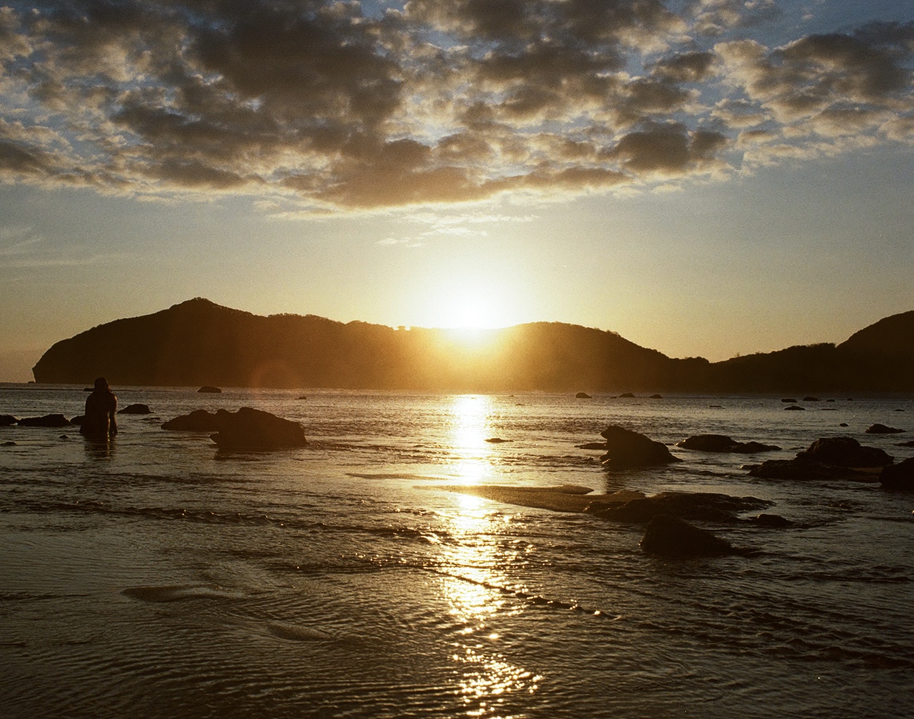 Person standing in shallow water as the sun sets behind silhouetted hills in Sumbawa