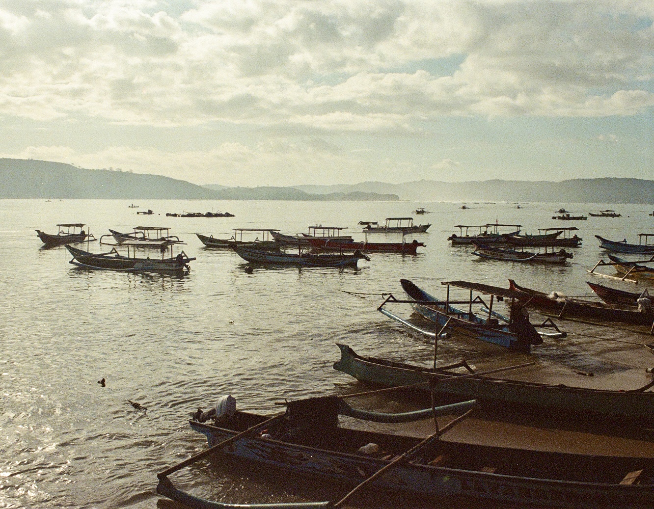Dozens of traditional fishing boats moored in a calm Indonesian harbour at dawn, misty hills behind