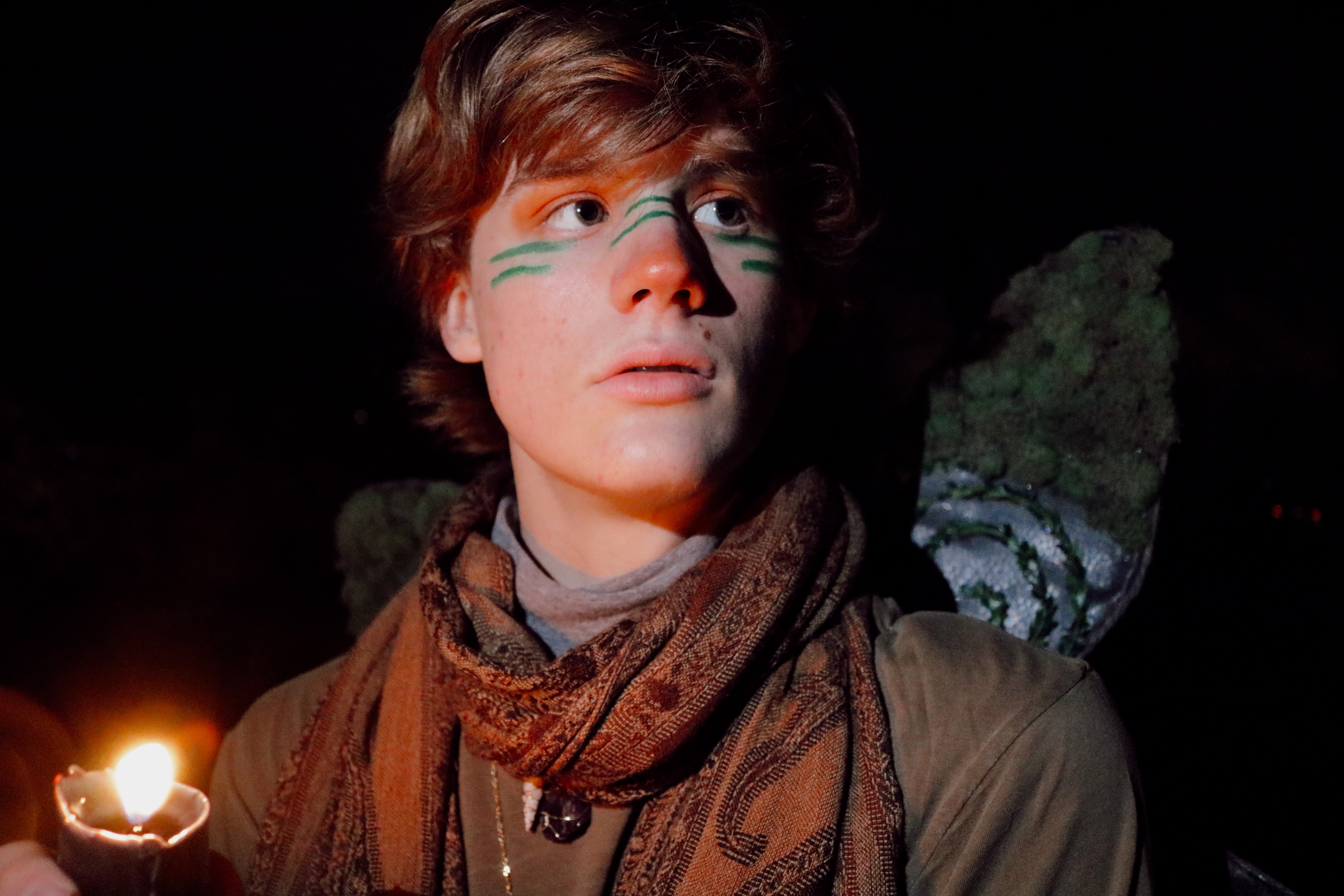 Young person with green face paint and moss wings holding a candle in darkness