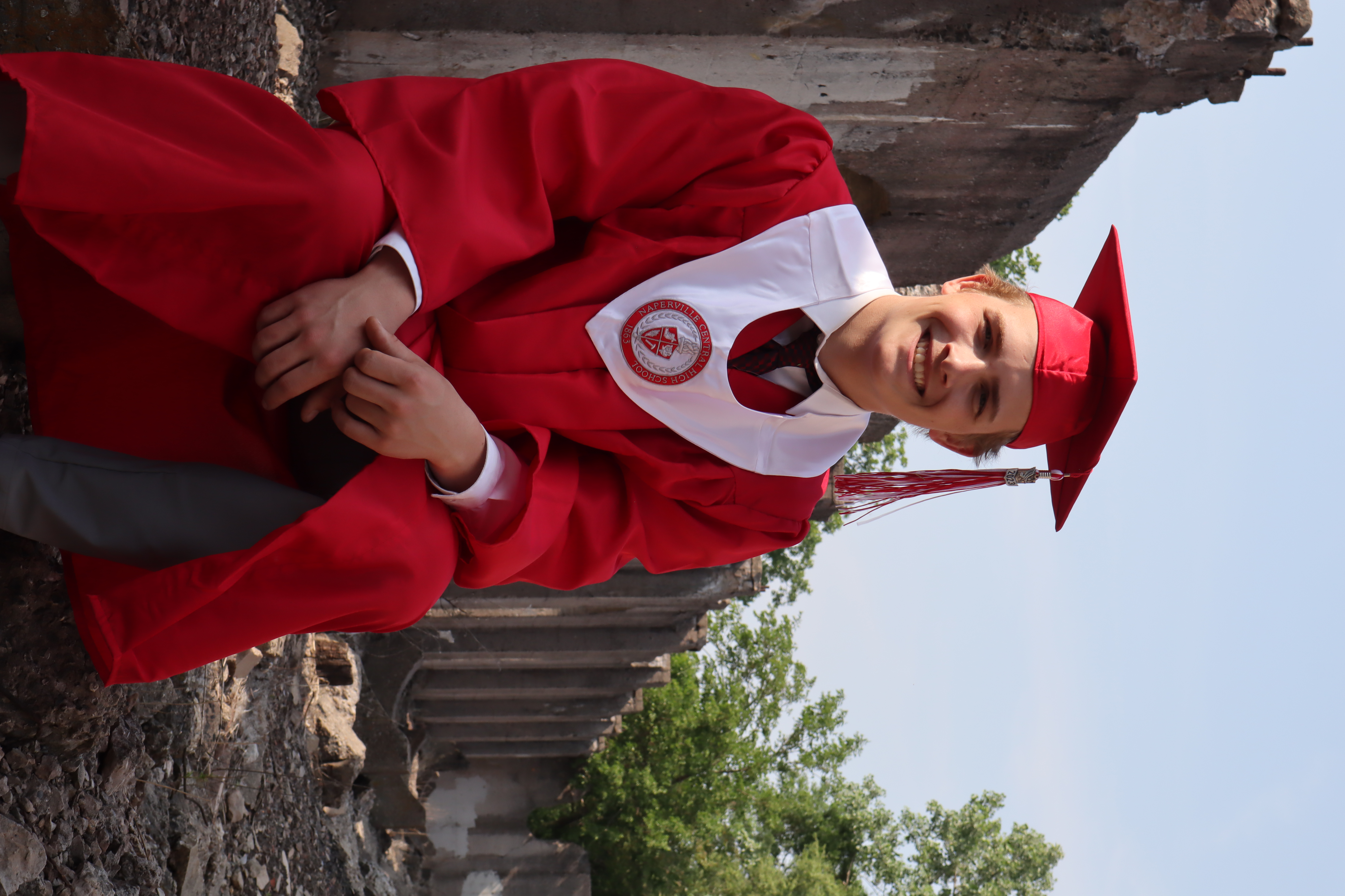 Mason in red cap and gown sitting among concrete ruins smiling