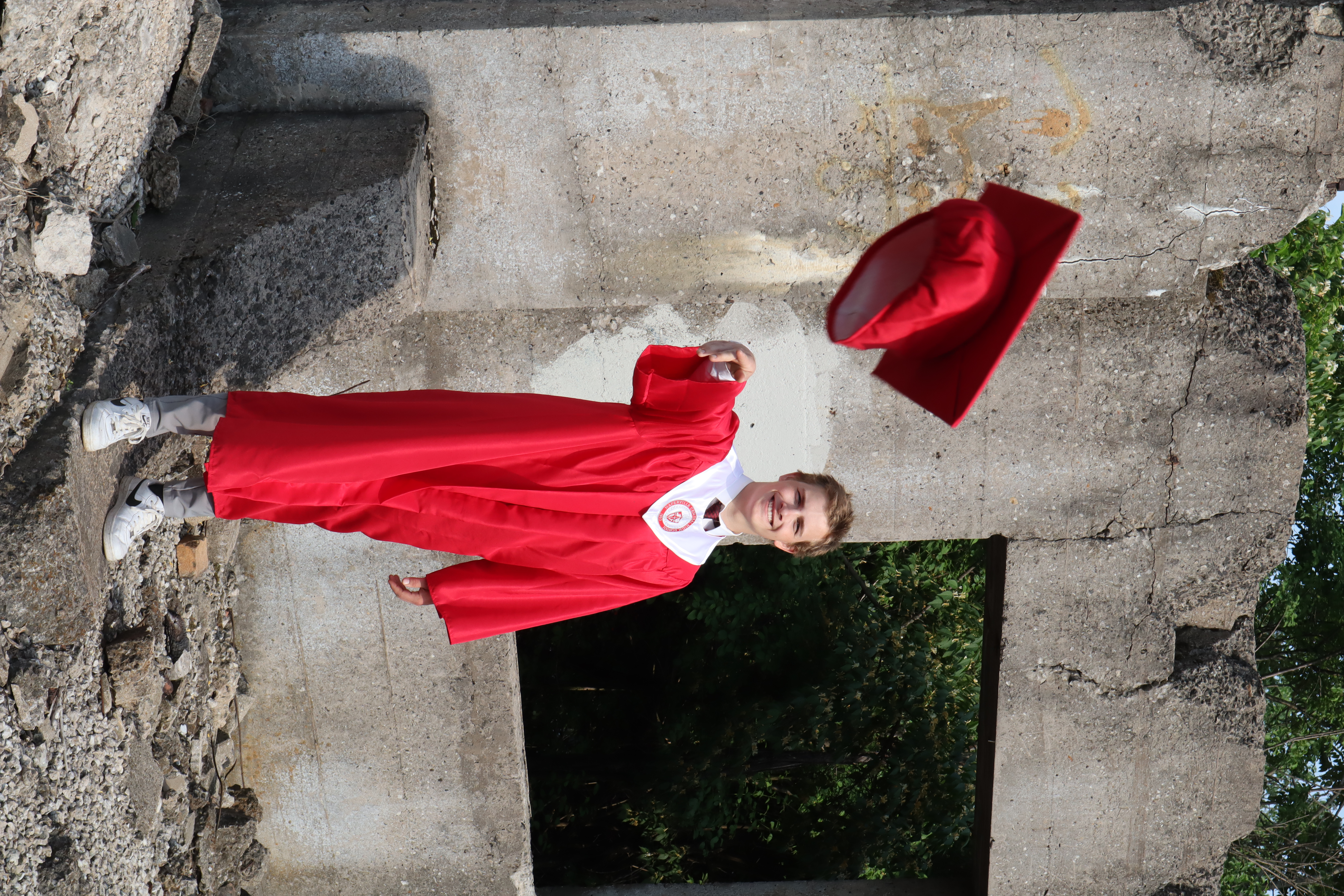 Mason in red gown tossing his graduation cap in the air at concrete ruins
