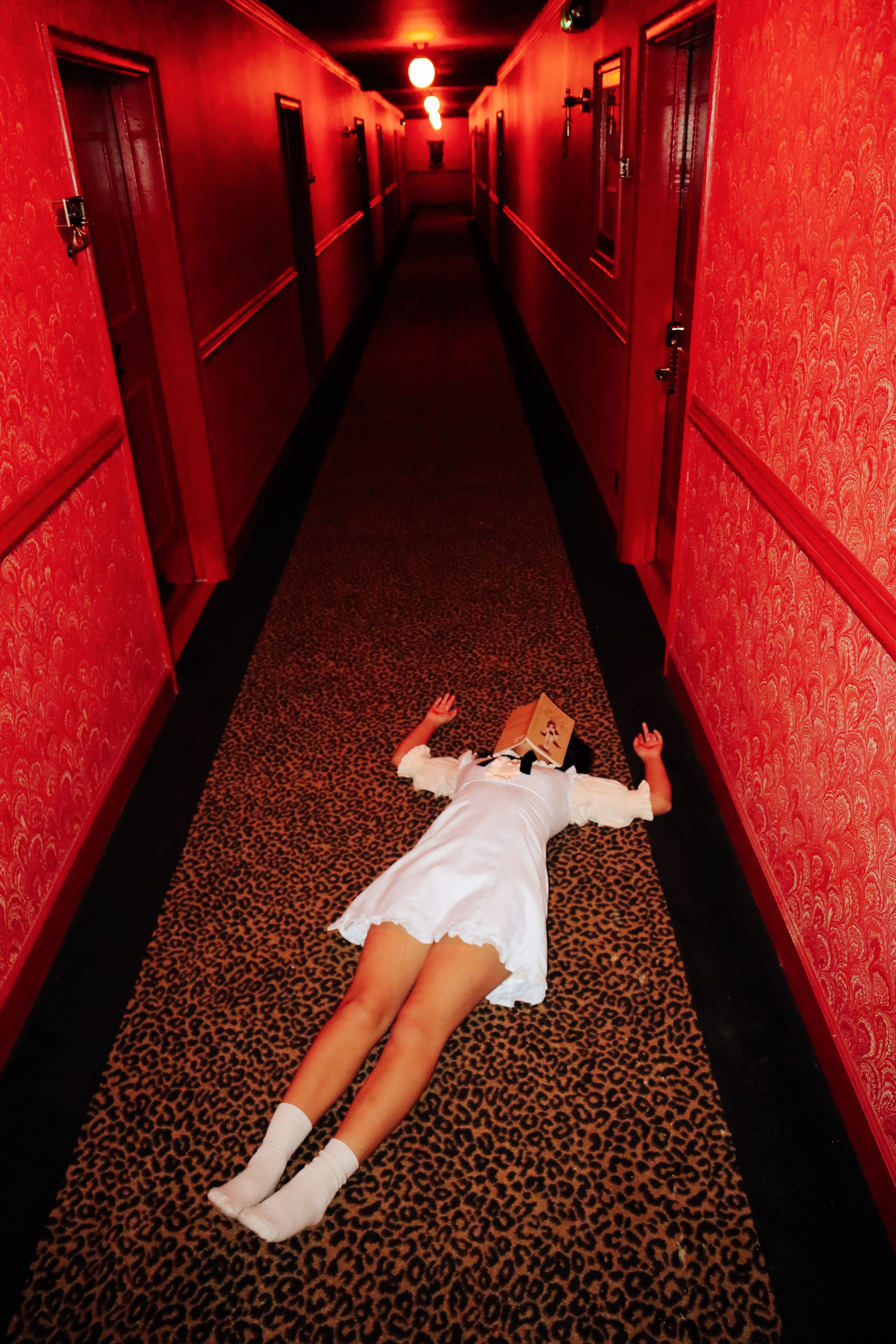 Woman in white dress lying on the floor of a red hallway with a box over her head
