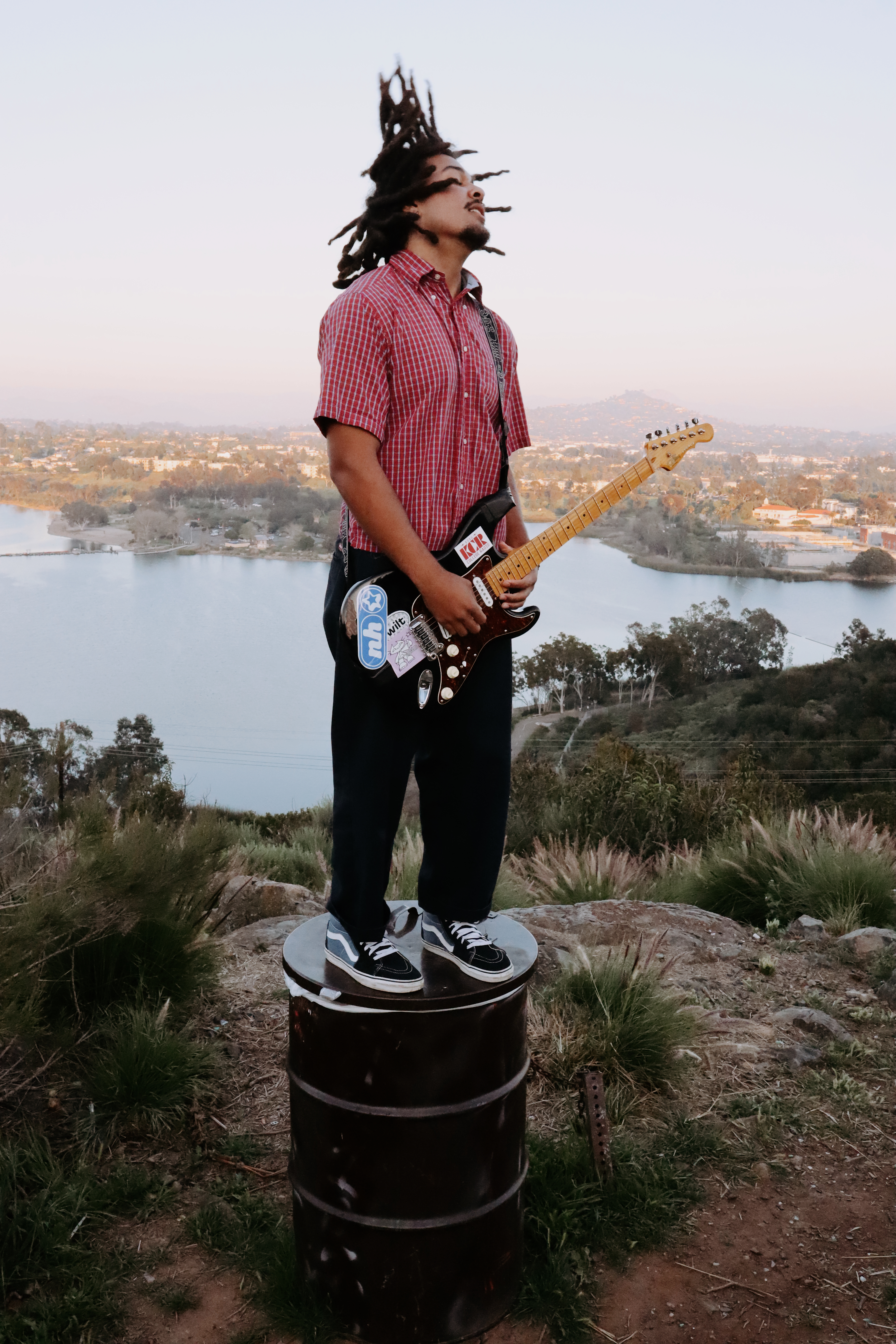 Guitarist standing on a barrel with dreadlocks flying, city below