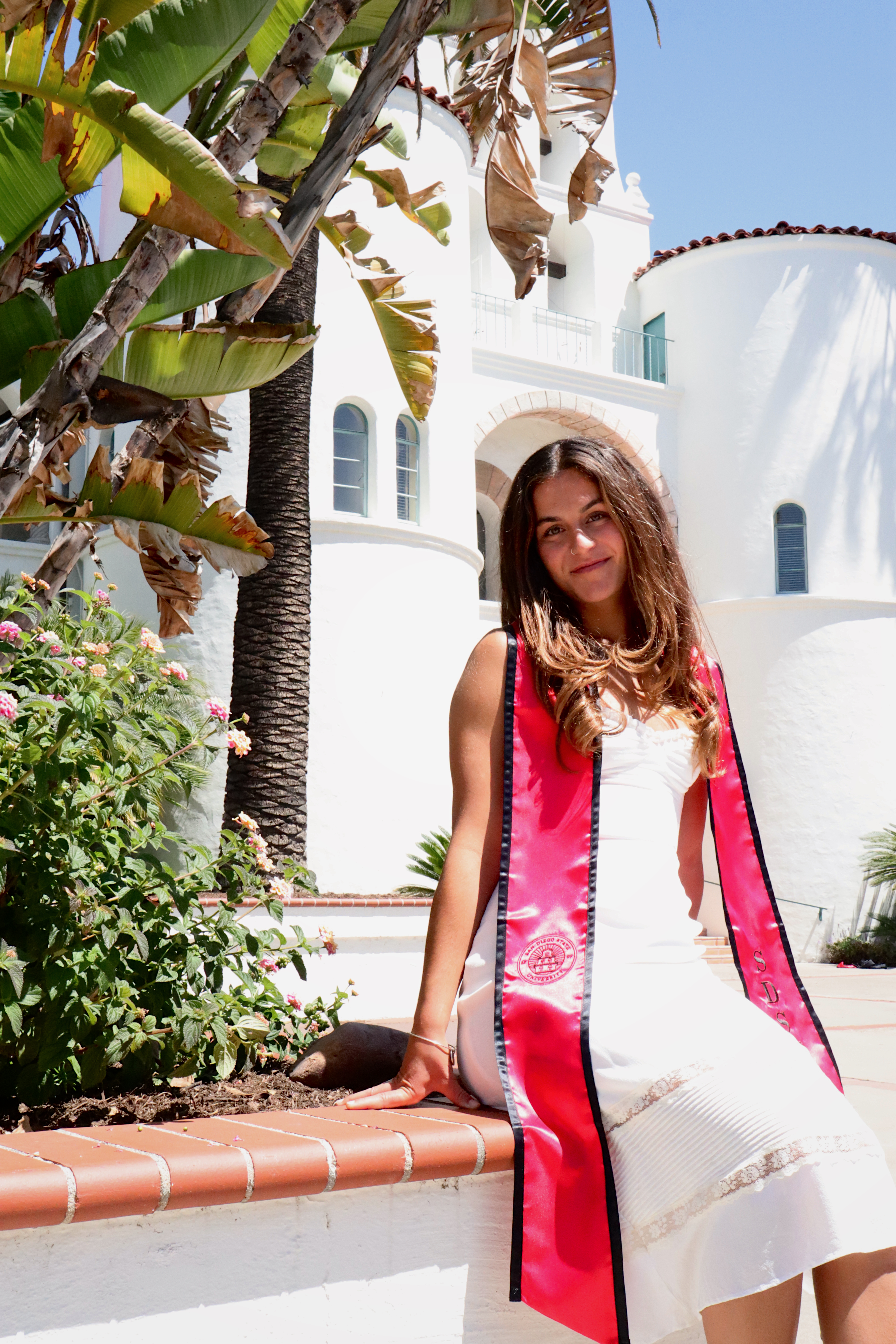 Kayden in SDSU graduation sash sitting on a ledge in front of the white campus building