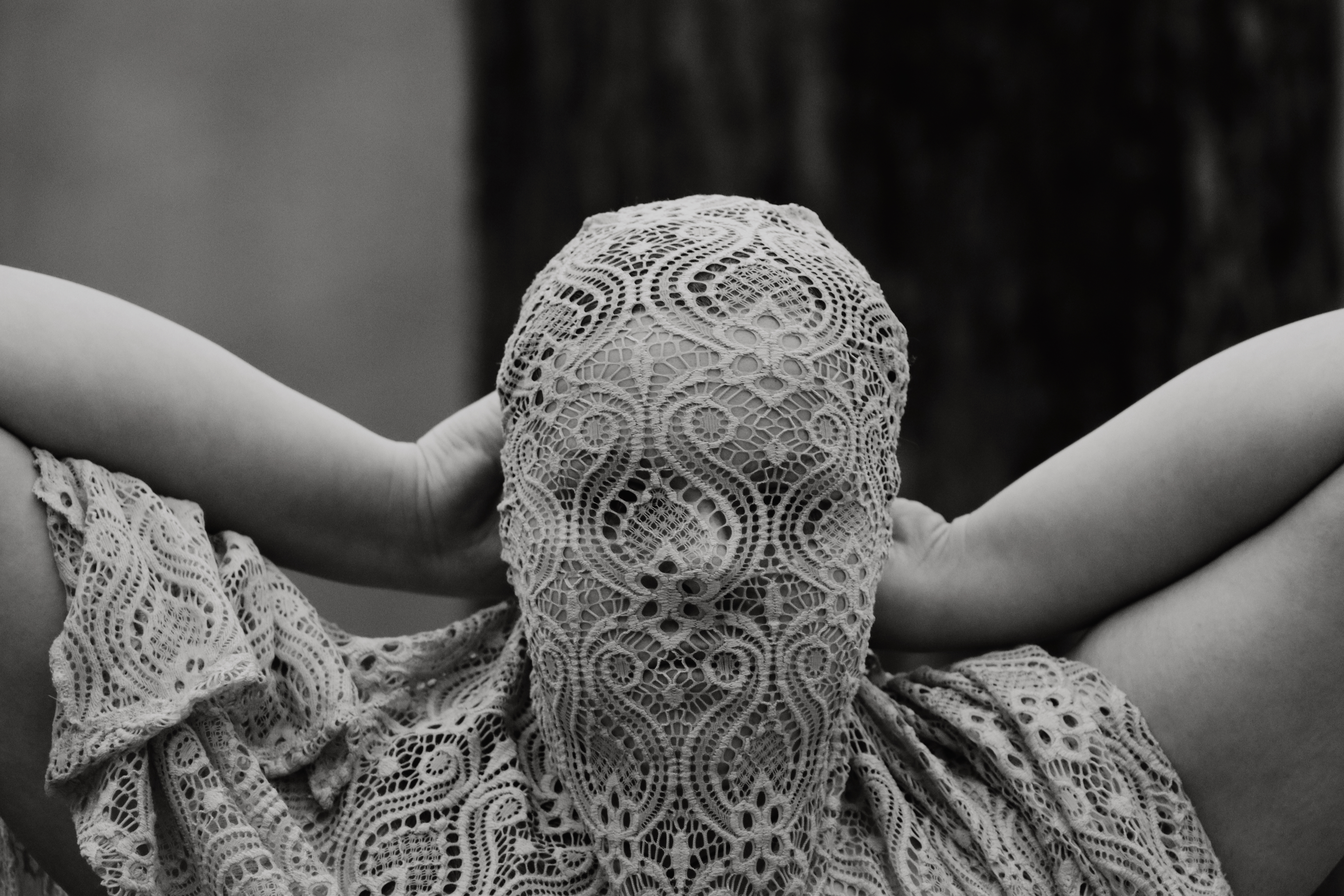 Person with lace fabric covering their face, hands raised behind head, black and white
