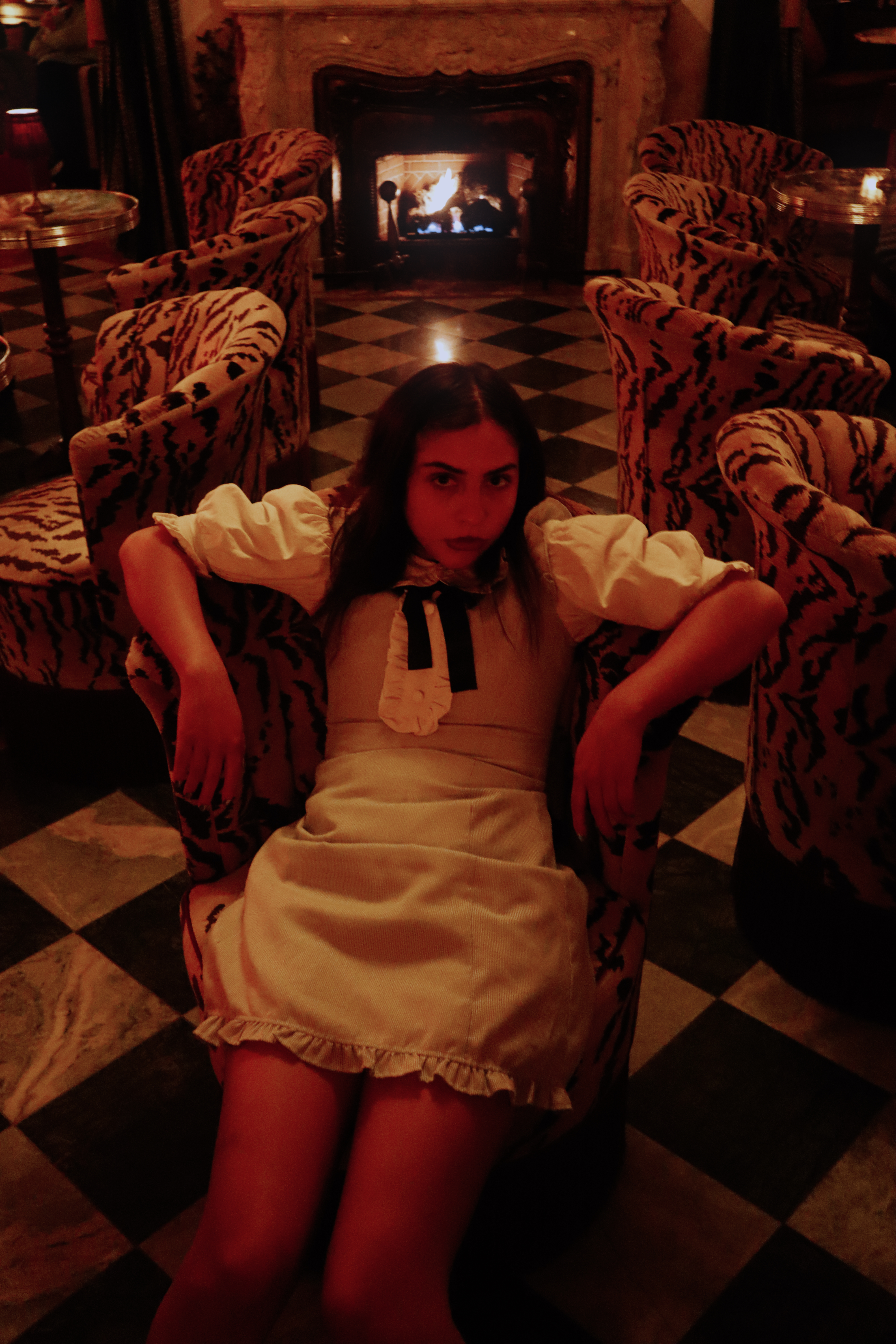 Woman in white dress sitting on checkered floor between tiger print chairs by a fireplace