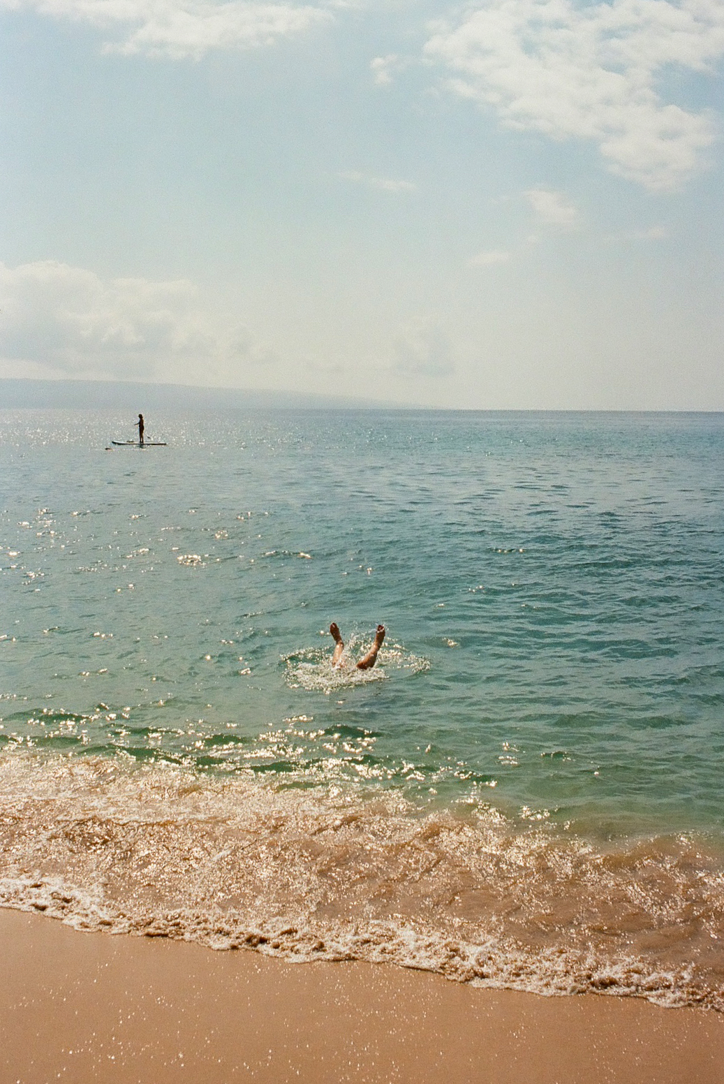 Person diving into turquoise Hawaiian ocean with paddleboarder in the distance