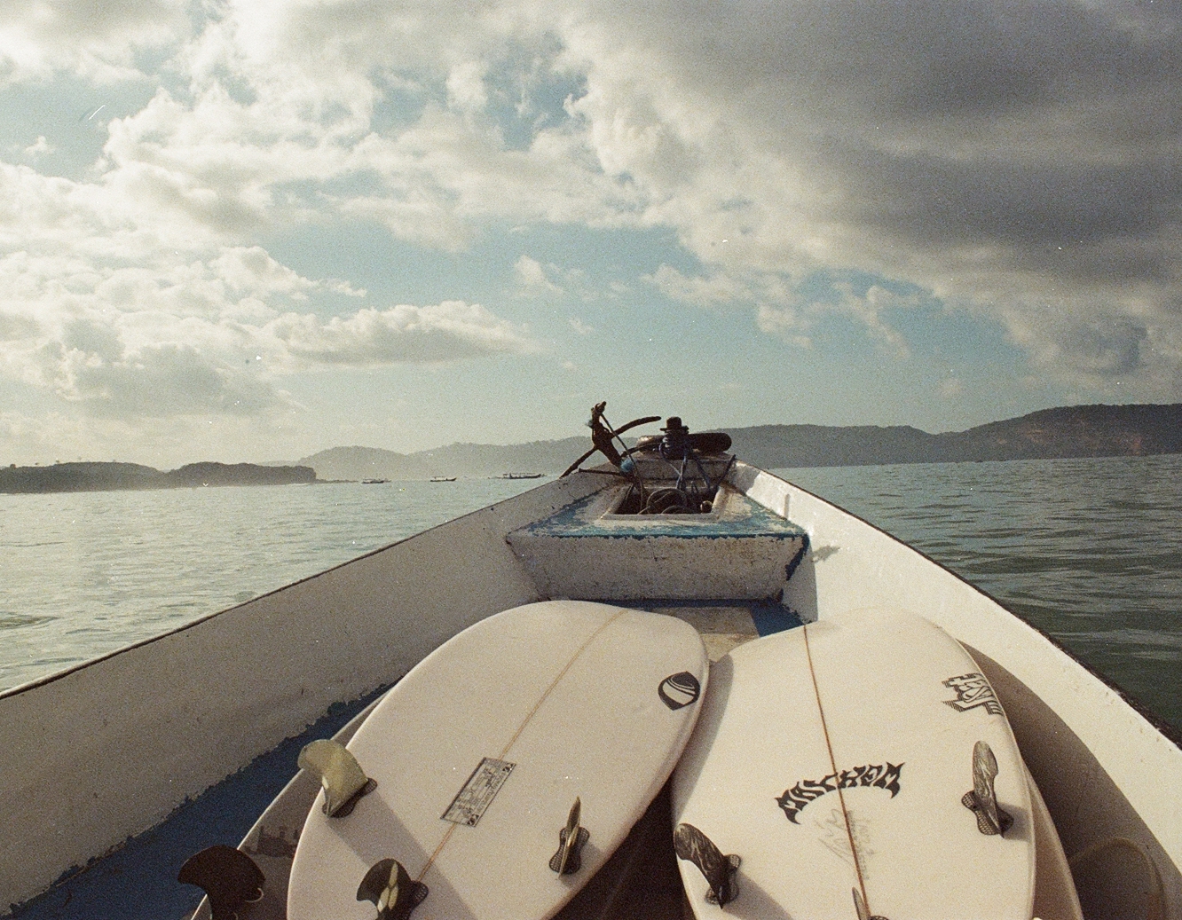 Surfboards stacked on the bow of a wooden boat crossing open water toward misty islands